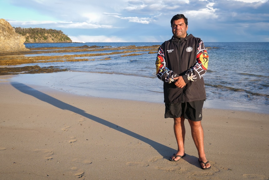 Man standing on a sunny beach, casting a long shadow on the sand behind him.