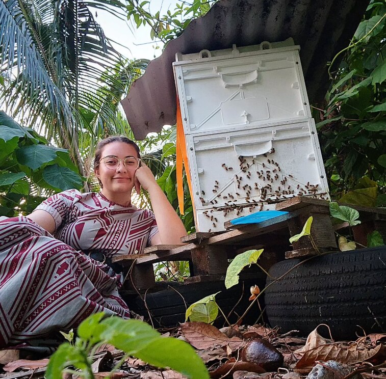 A girl in a Samoan dress sits next to a beehive in a green garden