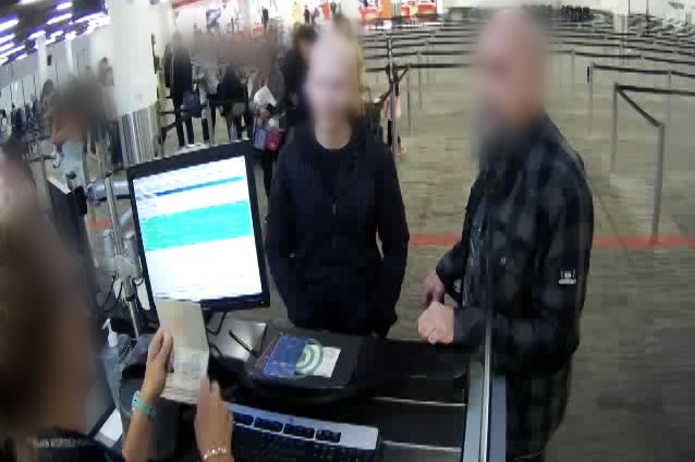 Two men stand at a customs desk at Perth Airport with their faces blurred as a customs officer checks a passport.