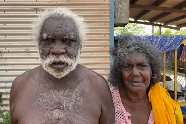 An elderly Indigenous man and woman standing side-by-side out the front of their home.