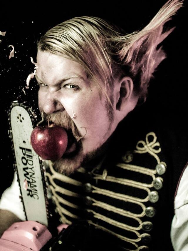 A circus performer holds an apple in his mouth and cuts it with a chainsaw.