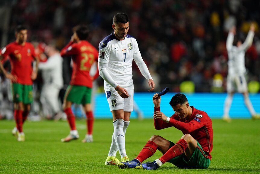 An upset football superstar Cristiano Ronaldo sits on the ground as an opposing player comes over to shake hands after a match. 