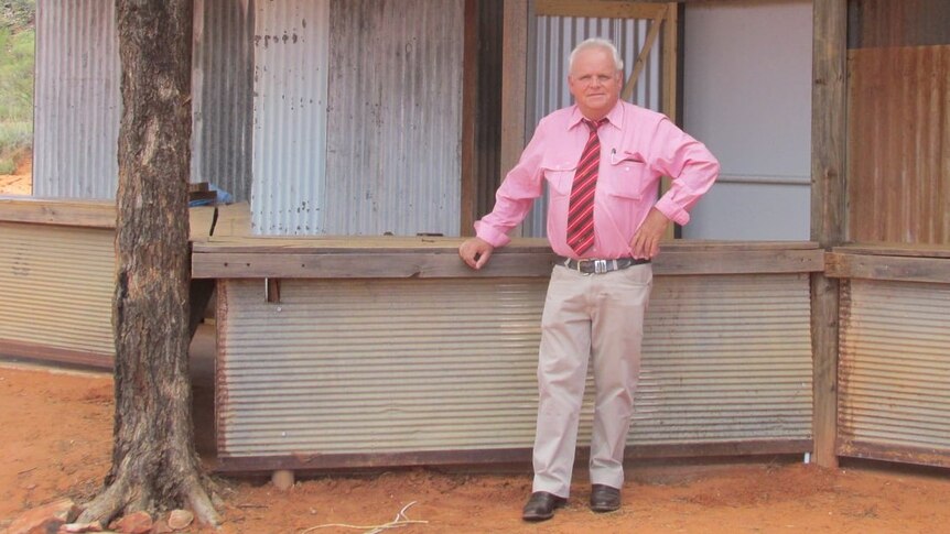 a man in a pink shirt standing in from of an old shed.