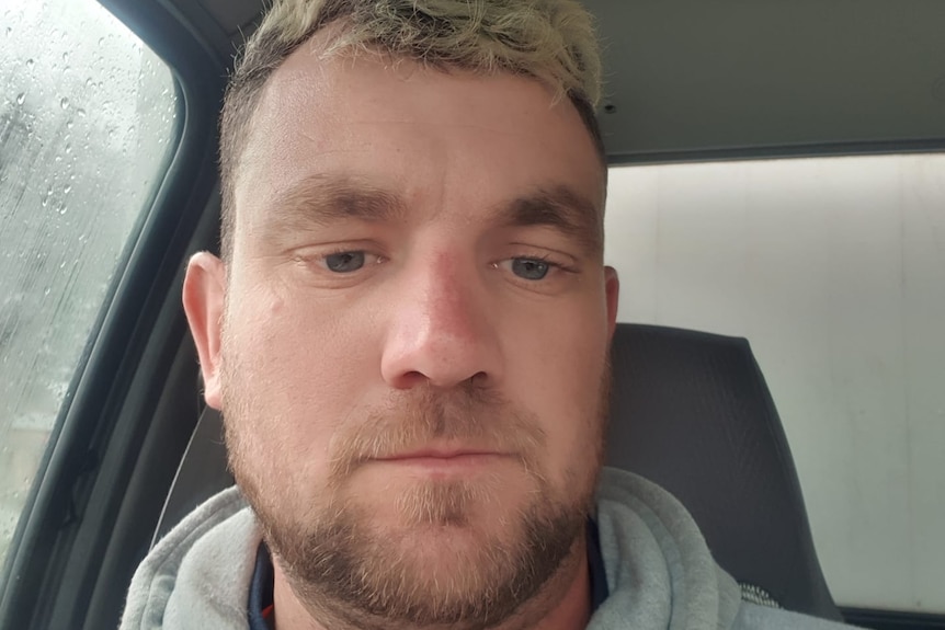 Un selfie d'un homme blond foncé avec une barbe courte et un sweat à capuche gris alors qu'il est assis dans une voiture