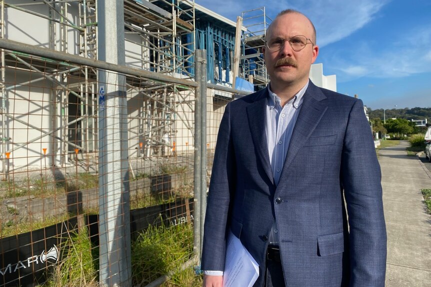 A balding man in glasses near a building site. He's wearing a navy suit with pale blue shirt and no tie, and carrying a papers.