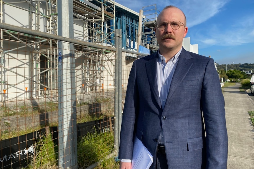 A balding man in glasses near a building site. He's wearing a navy suit with pale blue shirt and no tie, and carrying a papers.