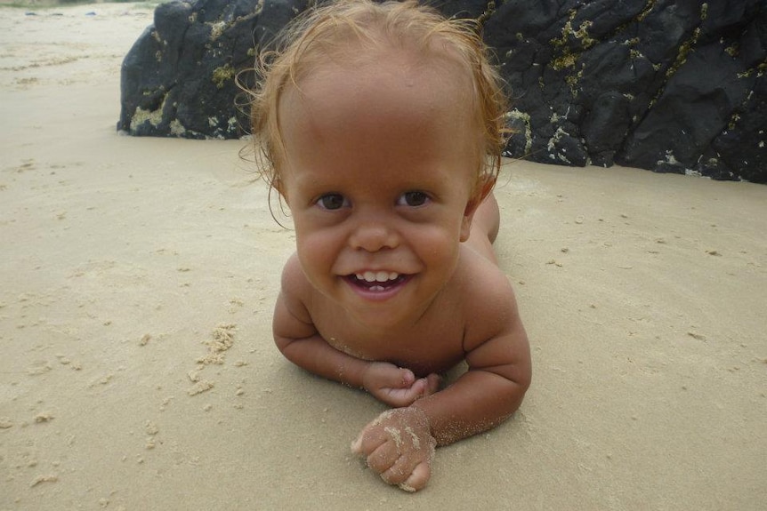 A young toddler boy smiles at the camera as he lays on his stomach at the beach