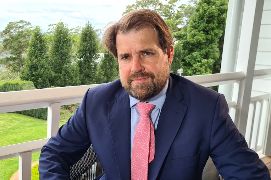 A man in a suit and tie is photographed sitting on the verandah of a building.