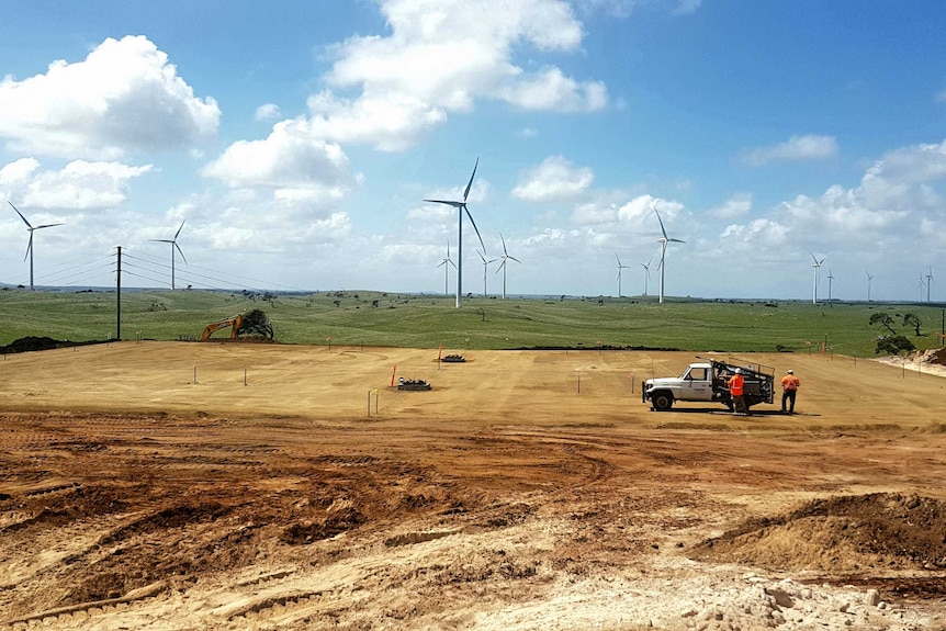 A flat area of dirt with grass and wind turbines in the background.