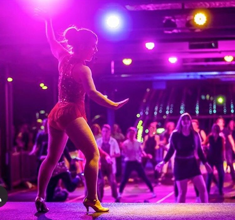 Neridah Waters stands in a leotard on a stage. Dancers are in the background behind her.