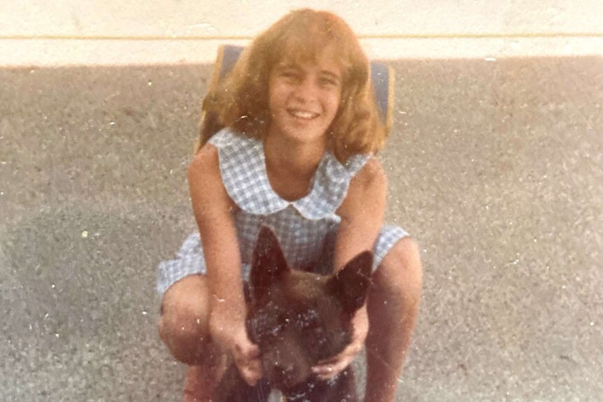 Old colour photograph of a school girl smiling at the camera, holding a kelpie by the mouth to make it smile