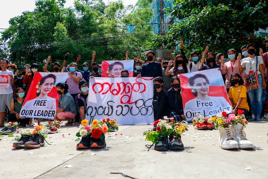 A group of people saluting the air with posters of Aung San Suu Kyi stand behind shoes filled with flowers.