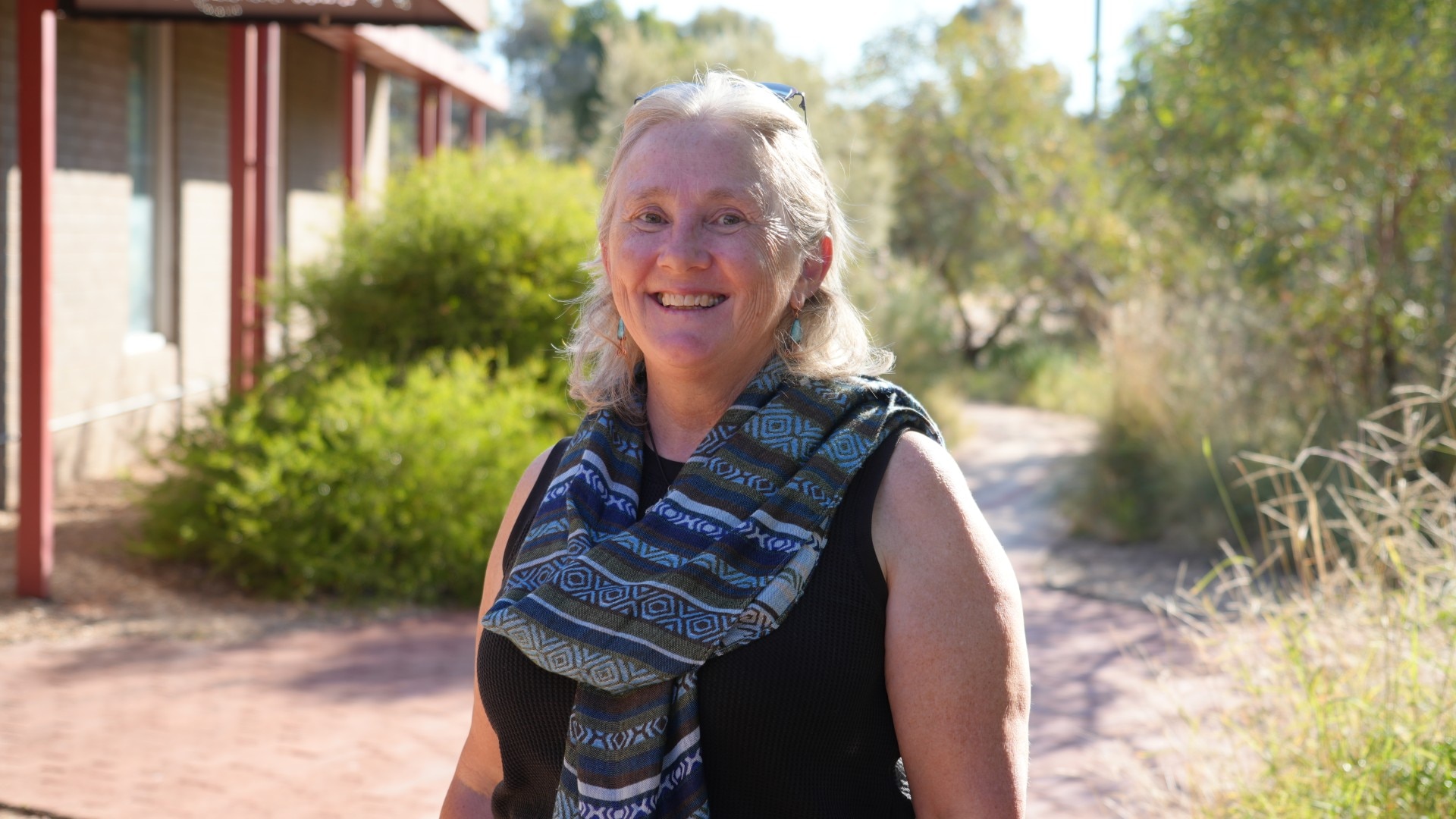 A woman wearing a blue patterned scarf stands in front of green bushes smiling
