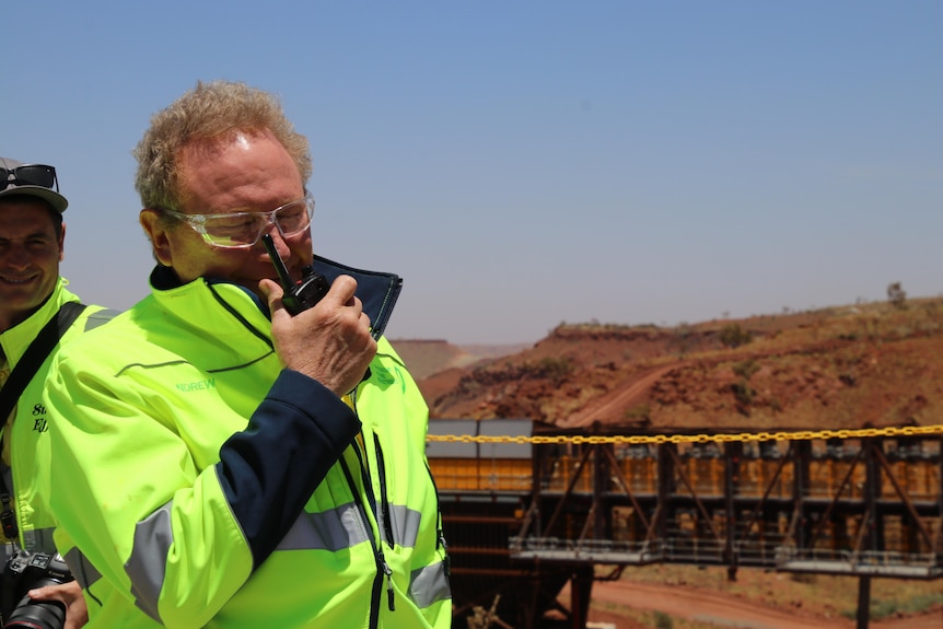 A man with a bright yellow jacket holds a walkie talkie in front of his mouth. He's standing at a mine site.