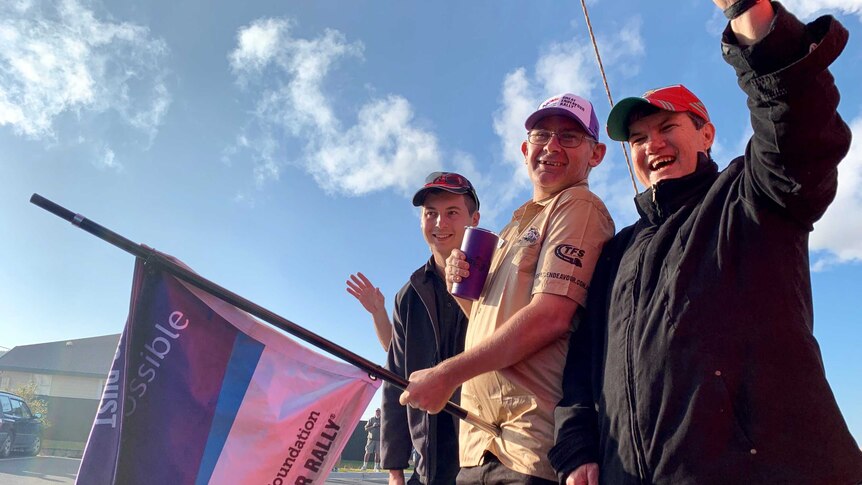 Two men and a woman stand smiling and waving a flag for the Endeavour Foundation.