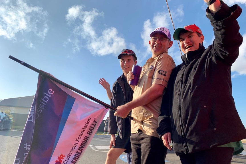 Two men and a woman stand smiling and waving a flag for the Endeavour Foundation.