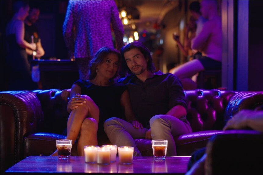 A man and a woman sit close together on a couch in a purple-lit bar. Lots of candles sit on a table in front of them.