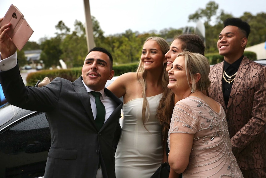 4 year 12 students, and a teacher, pose for a selfie in front of a limousine, wearing formal attire.