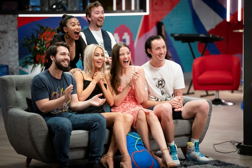 A group of young people sit on a couch with a few sitting behind it, they are smiling and some are clapping