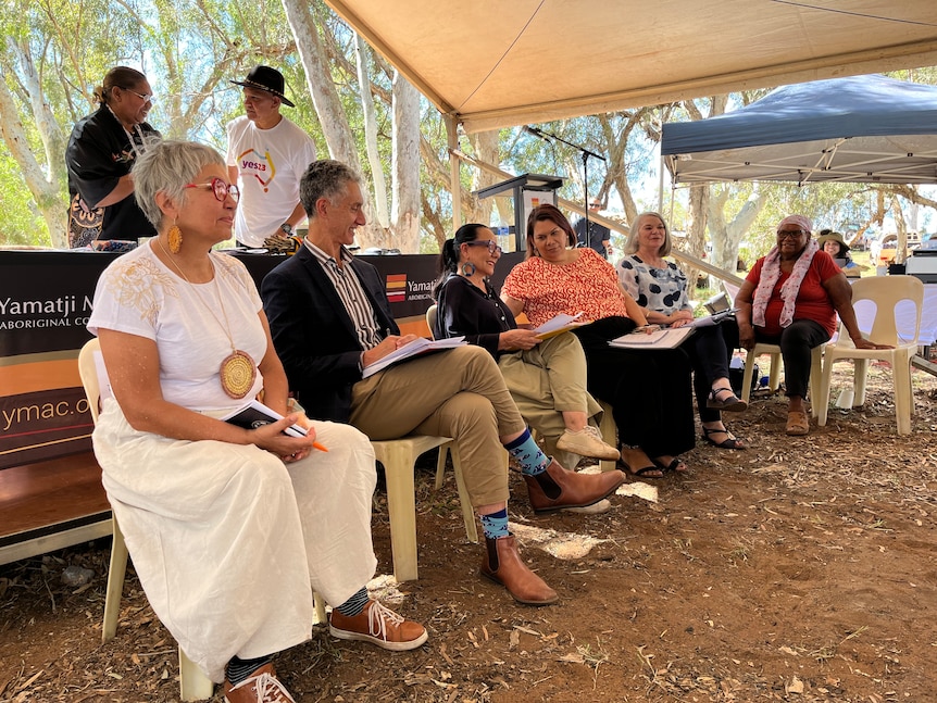 People sit in chairs under a marquee in the bush.