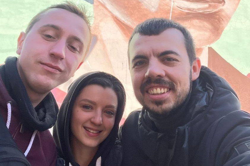 A man takes a selfie with his male and female friend. There is a mural on the wall behind. 