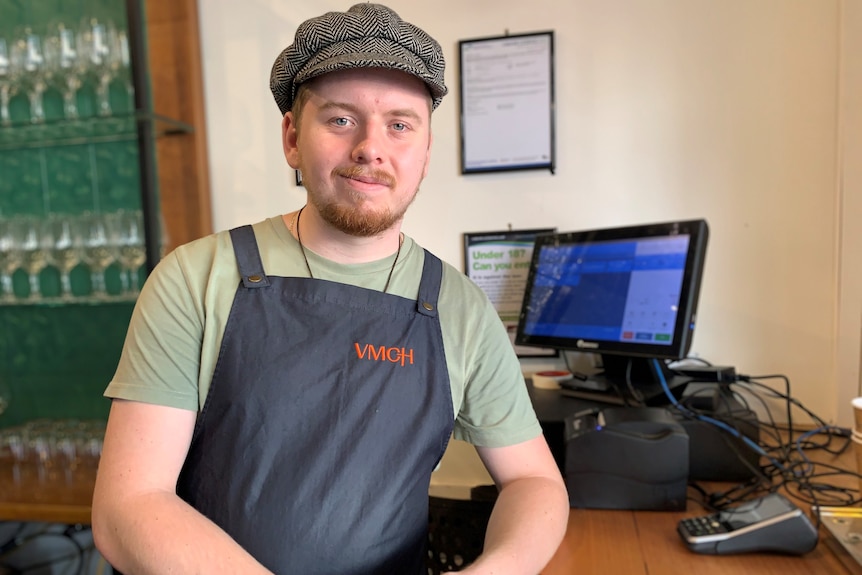 Trainee Zach Maskell,  wearing a newsboy cap,  leans on a counter in front of a cash register. 