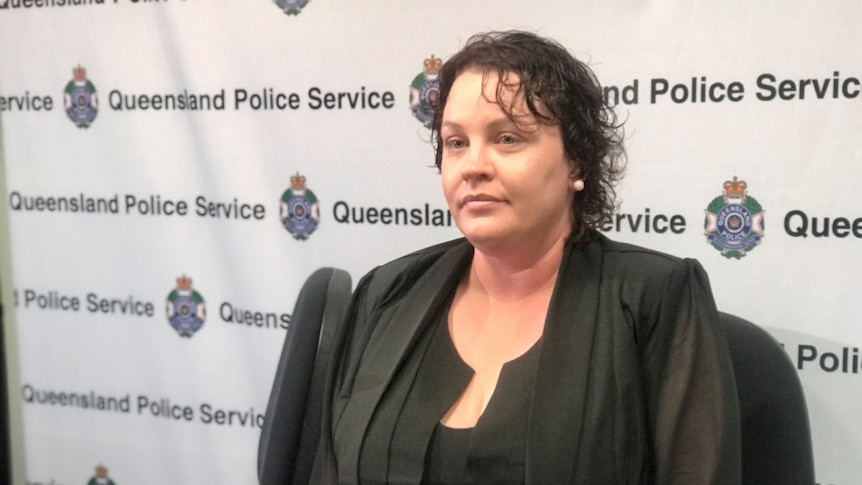 a woman in black clothing sits in front of a queensland police banner