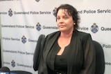 a woman in black clothing sits in front of a queensland police banner