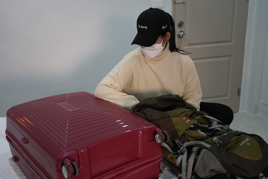 A young woman whose face is obscured by a black cap and white face mask sits on a bed next to a suitcase and backpack