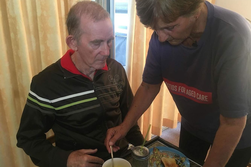 A woman stands beside a man sitting at a table and stirs soup for him