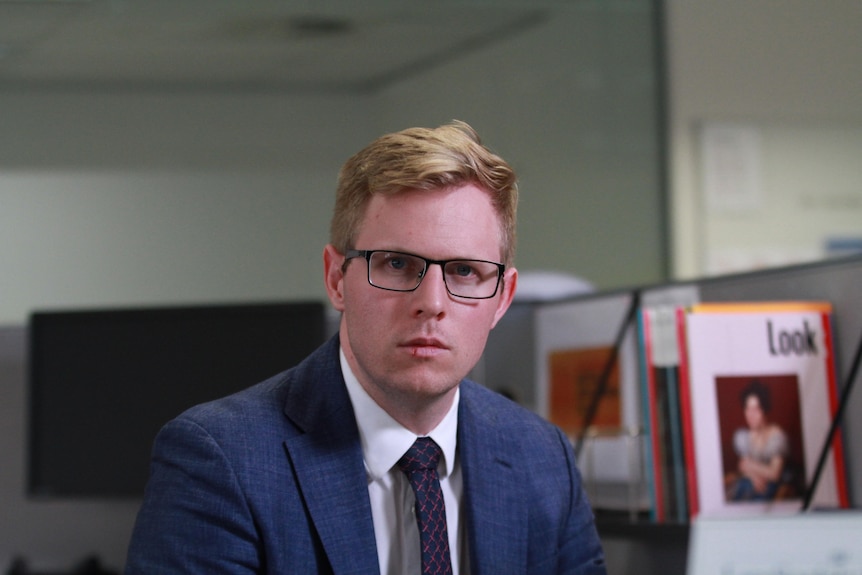 A man sits in an office, looking into camera with a serious expression.