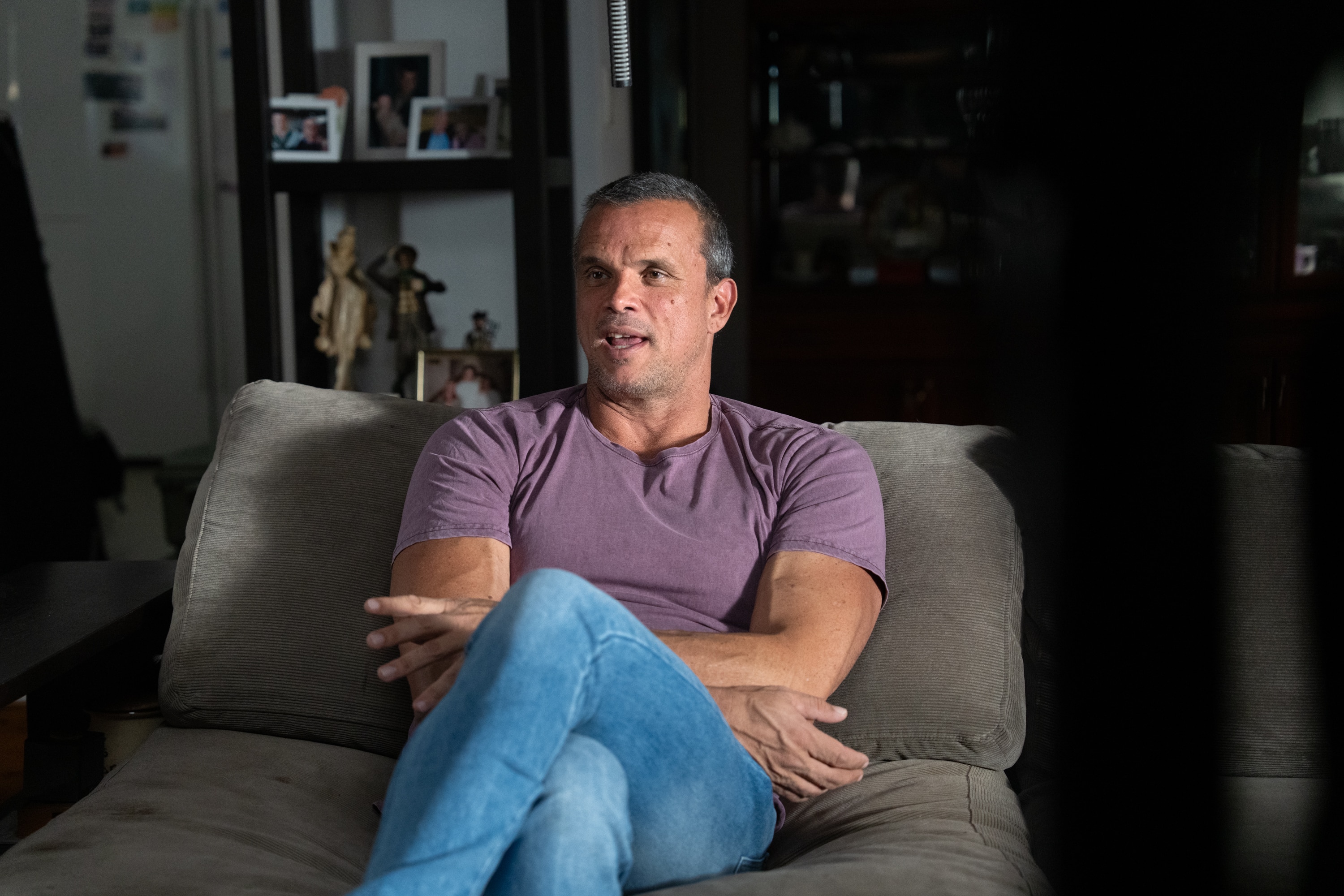 A man with short greying hair wearing a mauve tshirt and blue jeans sits on an arm chair in front of a bookcase.