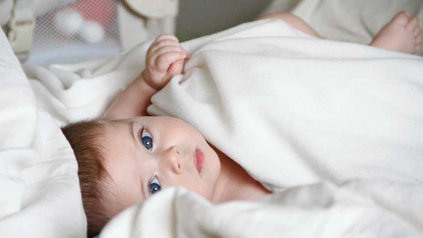 a blue eyed baby lies wrapped in a white blanket staring at the camera (and looking very sweet)