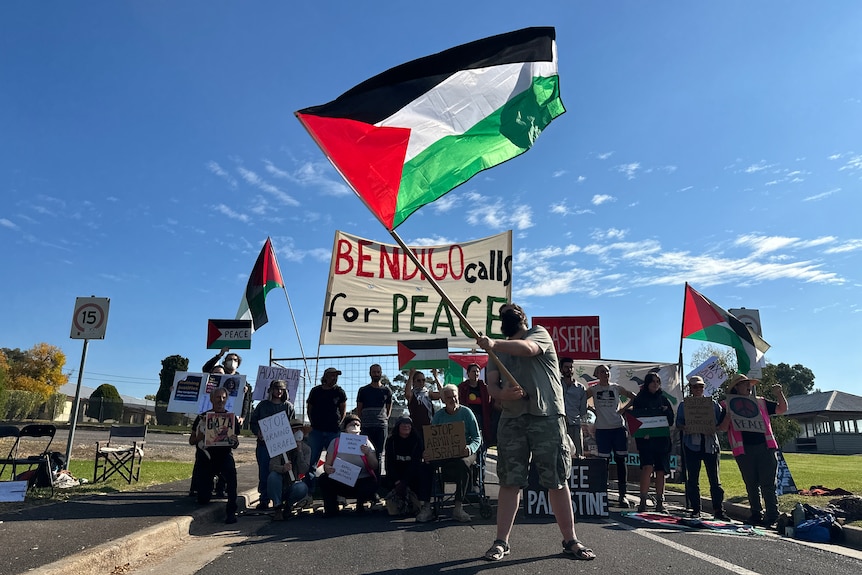 People holding flags and banners, gathered outside the gate to a factory.