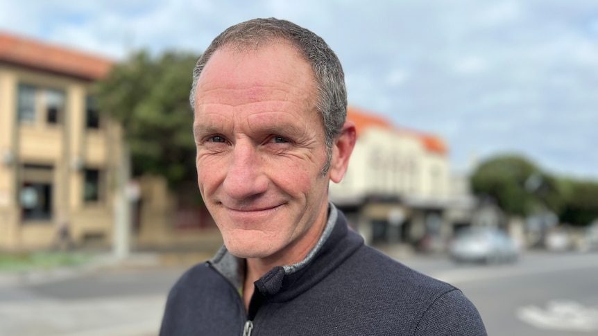 A middle-aged man with close-cropped grey hair and a black polo fleece standing in a regional town.