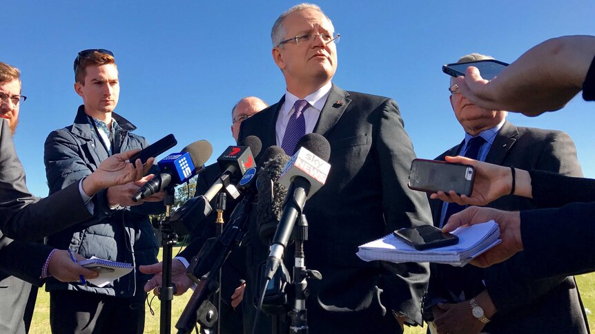 Scott Morrison stands in a press pack wearing a suit and speaking to reporters holding phones and microphones under a blue sky.