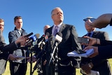 Scott Morrison stands in a press pack wearing a suit and speaking to reporters holding phones and microphones under a blue sky.