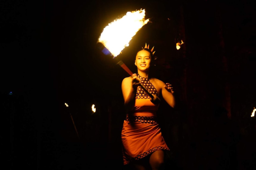 A woman in a orange dress swinging a large machete on fire.