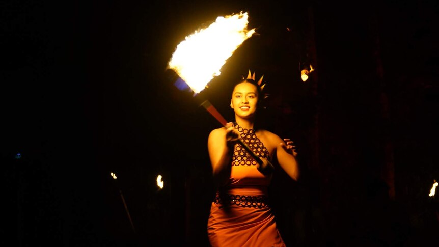 A woman in a orange dress swinging a large machete on fire.