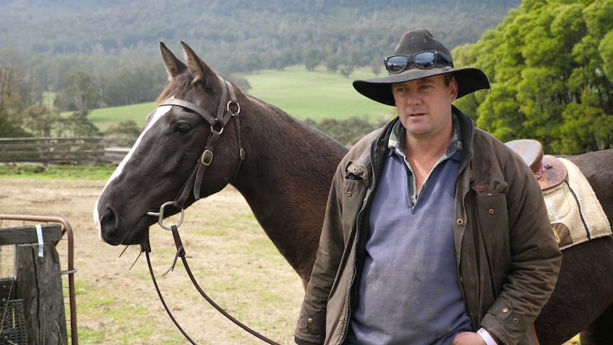 Jamie wearing a dri-z-a-bone and standing with his horse. Green paddocks and bush visible in the background.