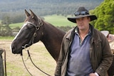 Jamie wearing a dri-z-a-bone and standing with his horse. Green paddocks and bush visible in the background.