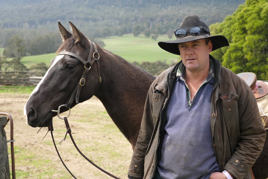 Jamie wearing a dri-z-a-bone and standing with his horse. Green paddocks and bush visible in the background.