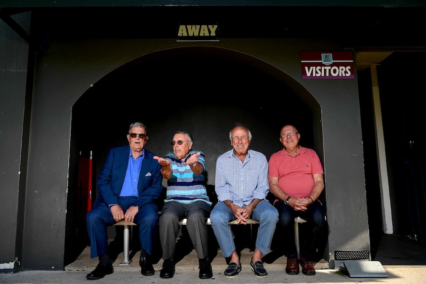 Four old men sit on a bench under the 'visitors' and 'away' signs.