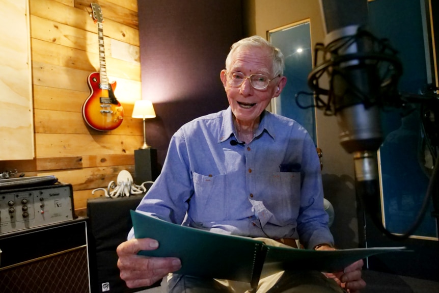 A man smiles at the camera. He is holding a folder of songs while sitting in a music studio.