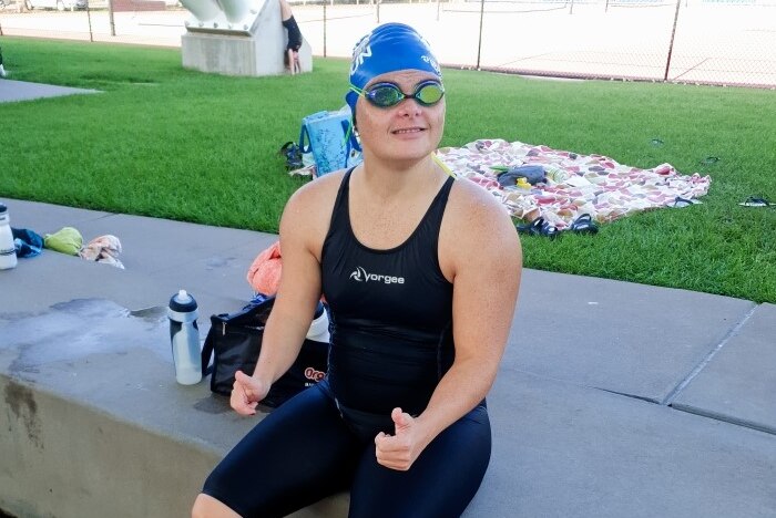 A woman with down syndrome sits on the side of a pool making a thumbs up gesture