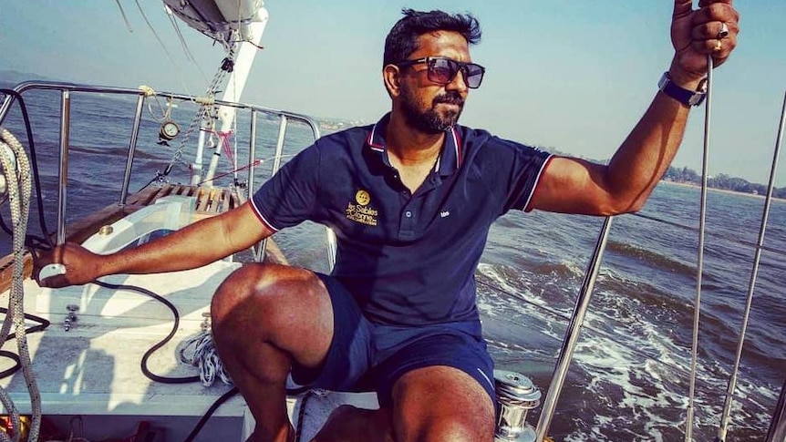 Solo sailor Abhilash Tomy sits on a yacht under a blue sky wearing shorts, a shirt and sunglasses.