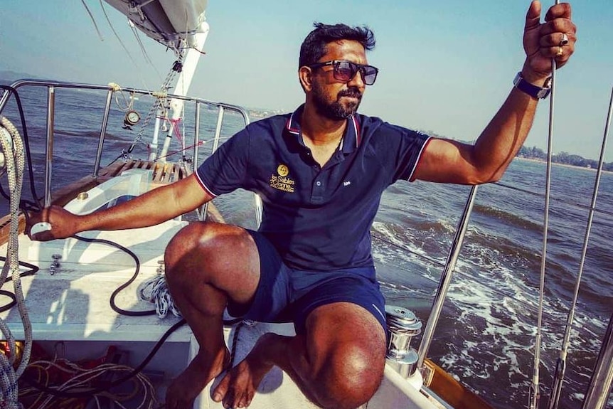 Solo sailor Abhilash Tomy sits on a yacht under a blue sky wearing shorts, a shirt and sunglasses.