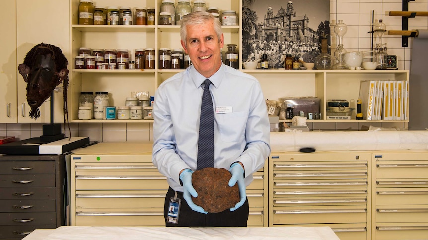 A man stands in a scientific laboratory, wearing gloves and holding a large meteorite.