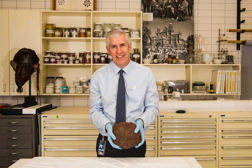 A man stands in a scientific laboratory, wearing gloves and holding a large meteorite.