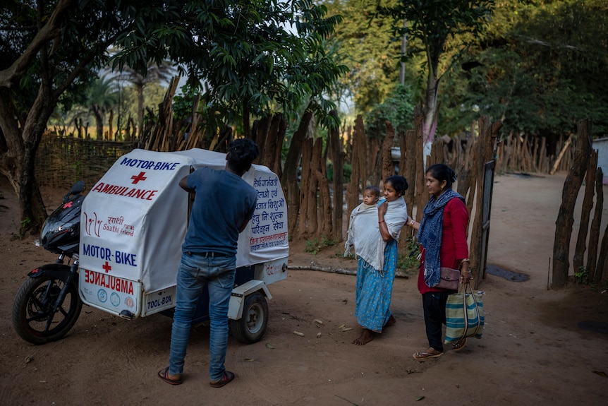 Two women, one holding a baby, walk towards a motorbike ambulance as a man holds it open.